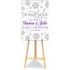 A Little Snowflake on the Way baby shower welcome poster board featuring silver snowflakes and purple lettering displayed on a wooden easel.